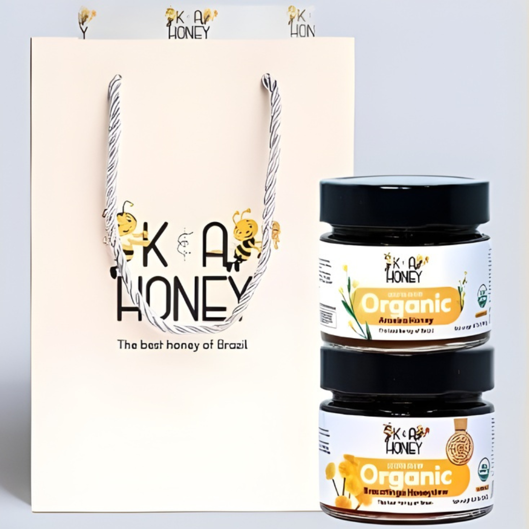 Cream-colored gift bag with rope handles featuring cartoon bees and 'K A HONEY – The best honey of Brazil'; includes two stacked jars labeled 'Organic Amazon Honey' and 'Organic Brazilian Honeydew' with white labels, yellow accents, floral illustrations, and organic certification.